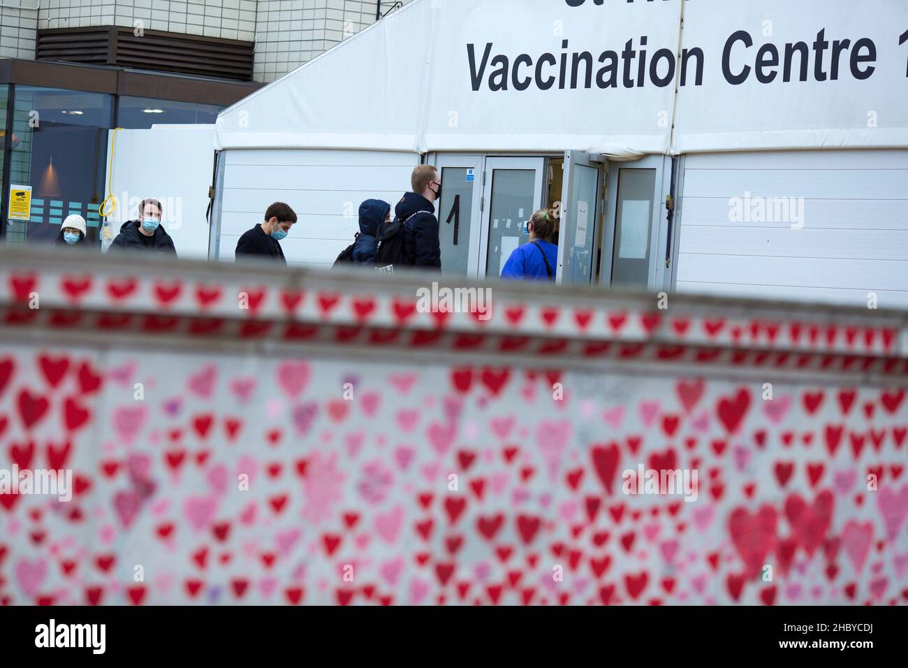 People are seen queuing towards a vaccination centre at St Thomas’ Hospital behind red hearts painted on the National Covid Memorial Wall in London. Stock Photo