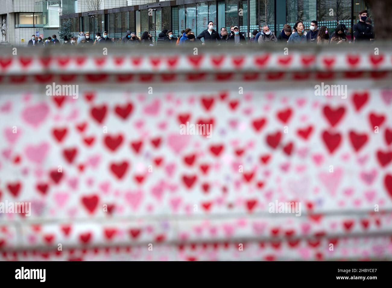 People are seen queuing towards a vaccination centre at St Thomas’ Hospital behind red hearts painted on the National Covid Memorial Wall in London. Stock Photo