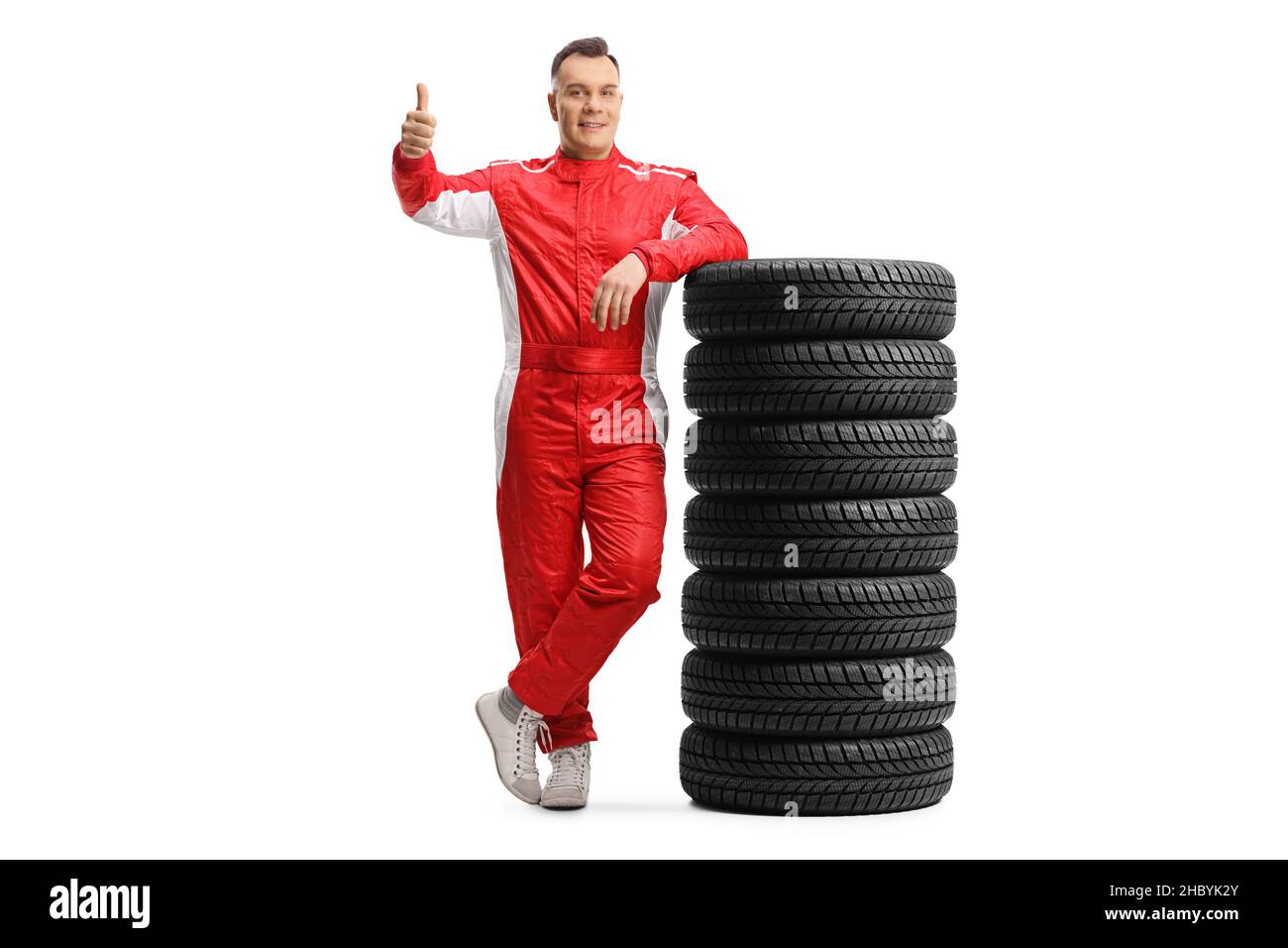 Full length portrait of a racer in a red suit leaning on a pile of tires and showing thumbs up isolated on white background Stock Photo