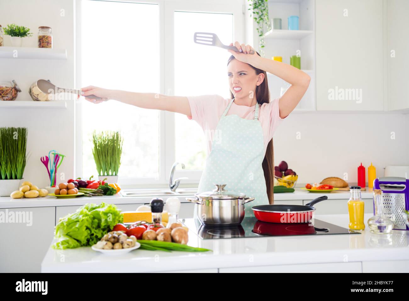 Portrait of attractive playful girl cooking tasty meal gourment having fun fighting with spoon in light white home kitchen indoors Stock Photo