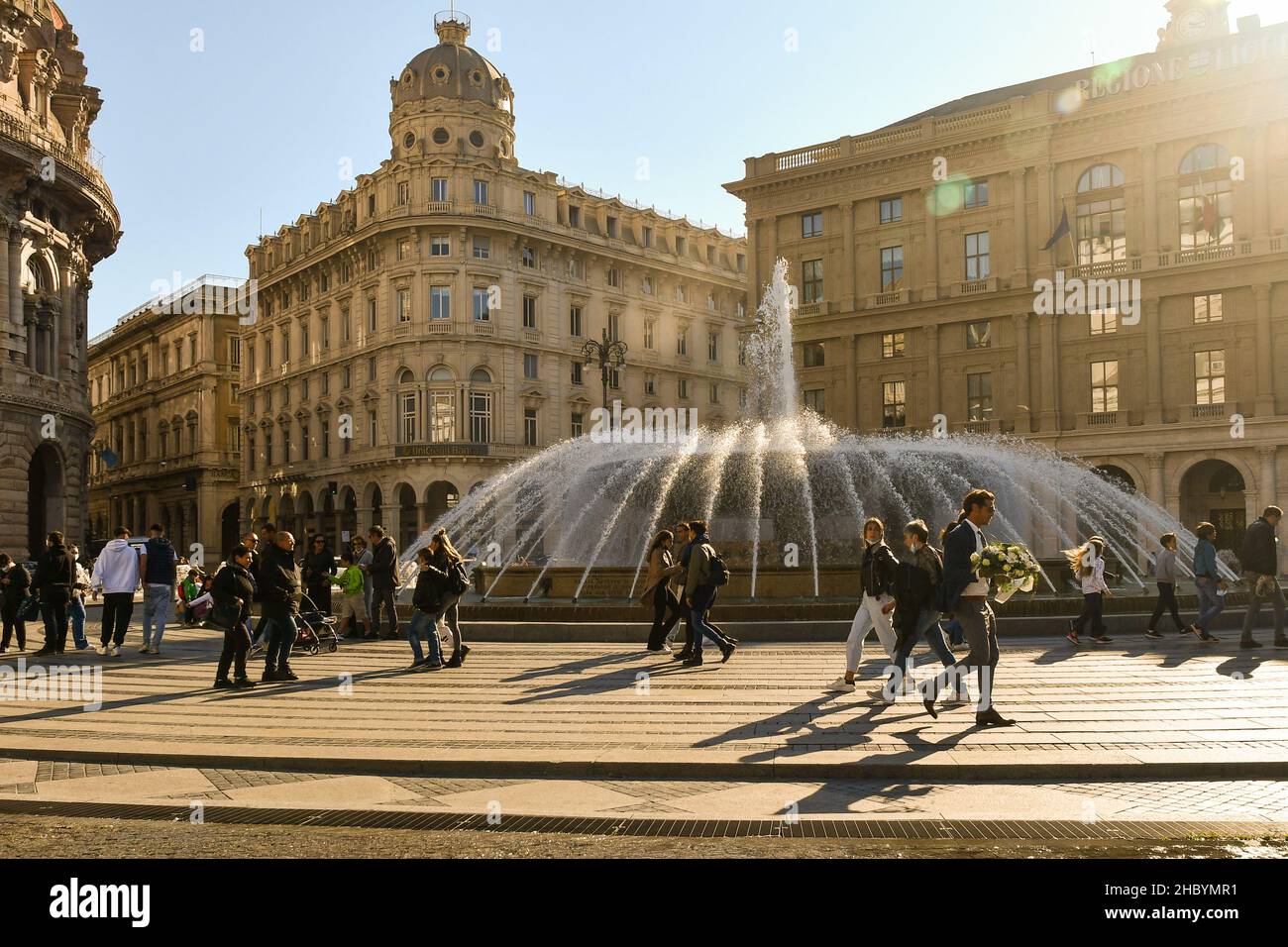 Backlight view of Piazza De Ferrari with people walking by the fountain and sun beams in a sunny autumn day, Genoa, Liguria, Italy Stock Photo