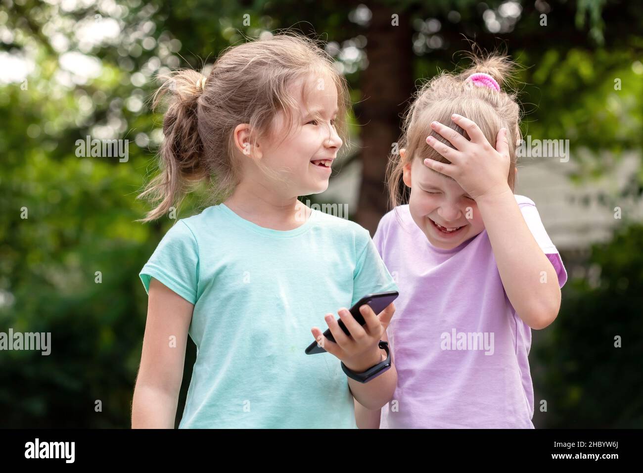 Two elementary school age children, girls, sisters siblings laughing, having fun using a smartphone, mobile phone, outdoors portrait, tech savvy kids Stock Photo