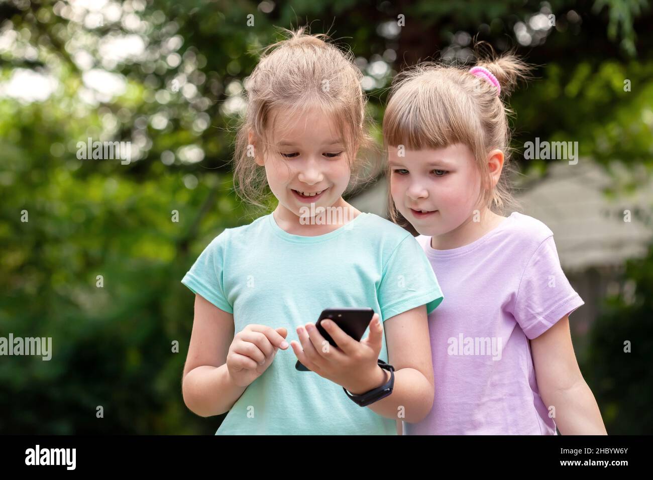 Two elementary school age kids girls, sisters, children laughing, having fun while using a smartphone, mobile phone, portrait, outside, tech savvy kid Stock Photo