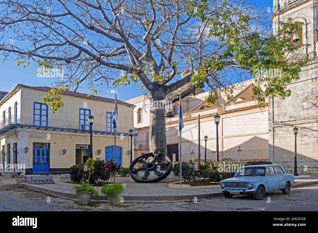 Kapok / ceiba tree (Ceiba pentandra), big old cogwheel and vintage car Lada VAZ-2101 at square in the city Matanzas on the island Cuba, Caribbean Stock Photo