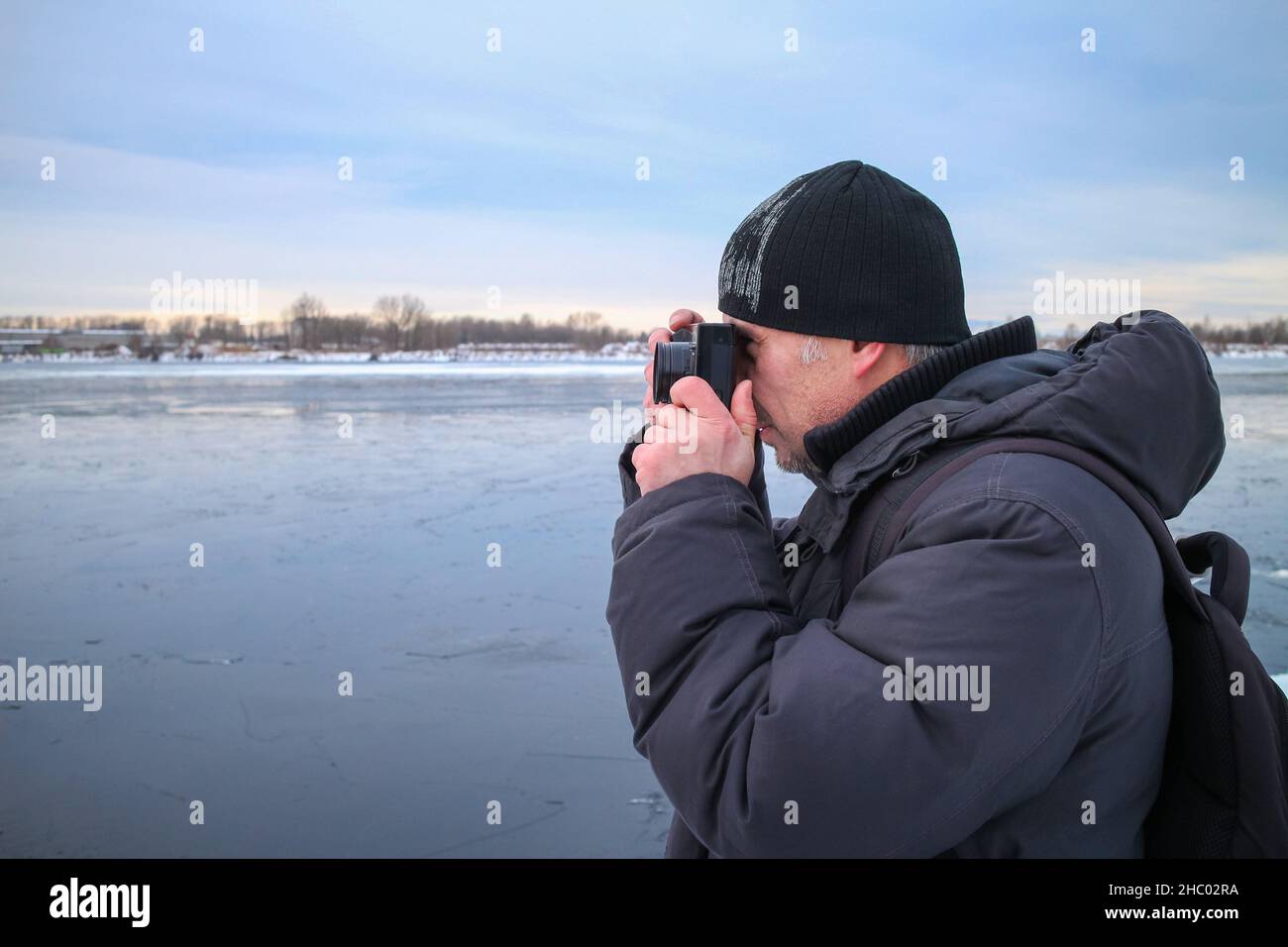 A man takes pictures with a film camera in winter in cold weather. The photographer looks into the viewfinder of the camera to photograph the winter l Stock Photo