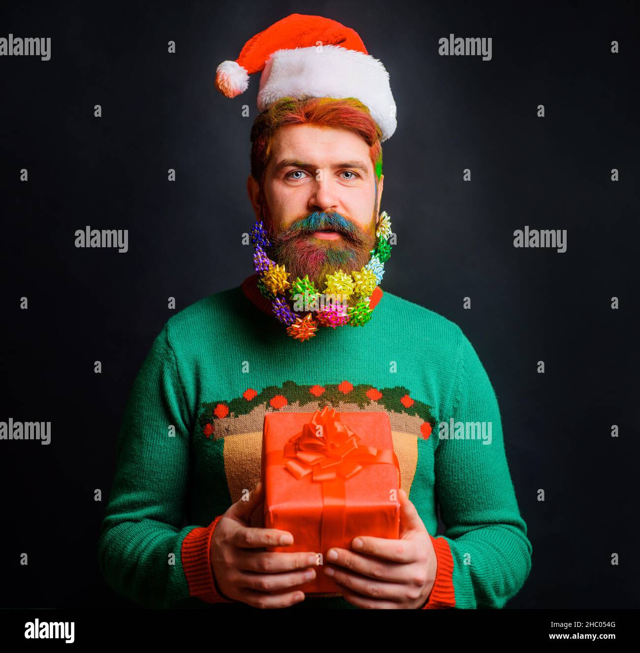 Bearded man in santa hat with Christmas gift. Hipster with decorated beard for New Year holiday. Stock Photo