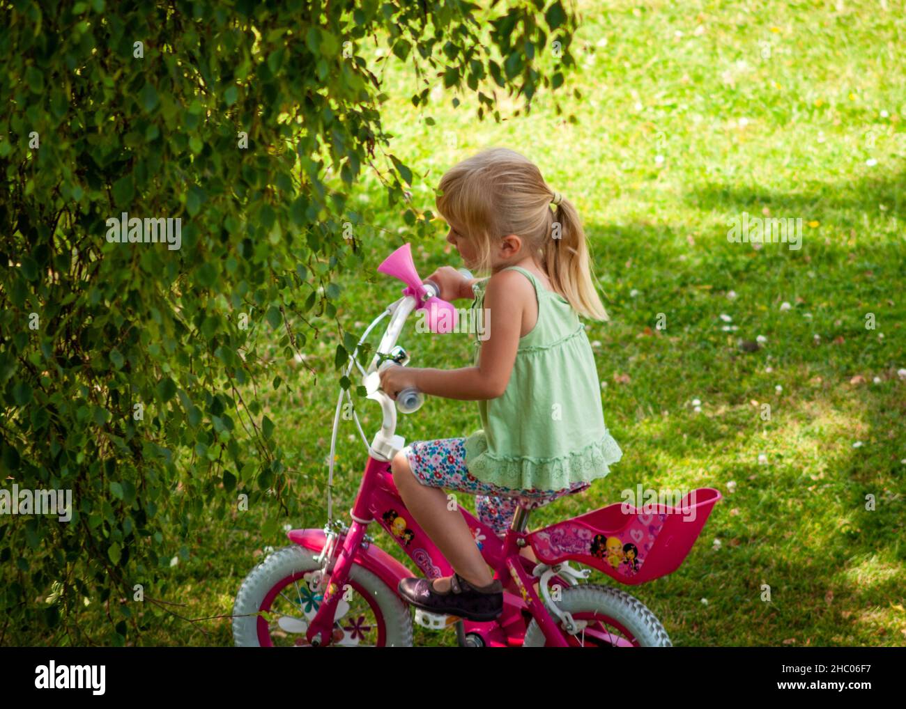 Little girl riding 12 inches pink girl's pedal bike with training wheels in open shade on green meadow backyard Stock Photo
