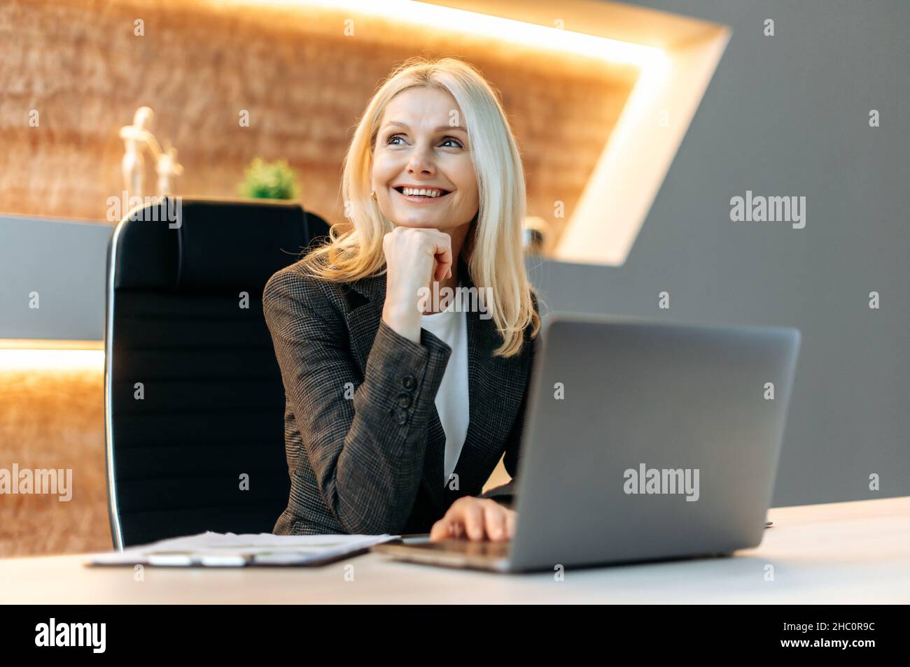Happy joyful successful blonde caucasian mature business woman, manager, finansist, sitting at table in formal clothes, taking break from work, looking pensively to side, dreaming about vacation Stock Photo