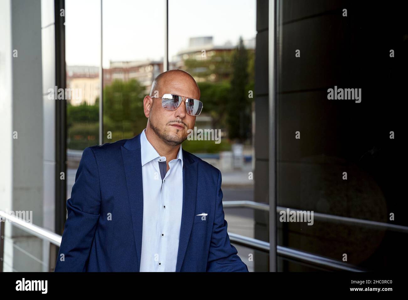 Bald adult male entrepreneur in smart casual clothes and sunglasses standing outside contemporary building with glass wall Stock Photo