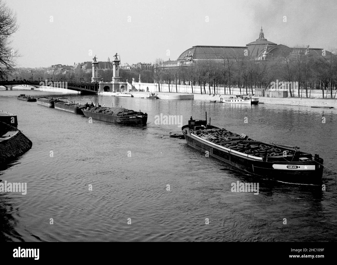 Paris River Seine with five cargo barges near Pont Alexandre III, 1945. The photographer stands on the Pont de la Concorde. The tow barge has already passed the field of vision. The lead barge in sight is loaded with tires. The Grand Palais is in the background. Stock Photo