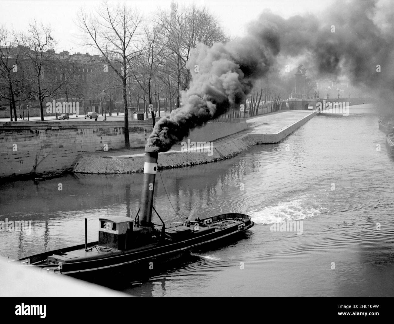 Paris Seine river smoky tow-barge at Pont de la Concorde, 1945. Smoke billows from a tow that is pulling out-of-view freight barges. Pont Alexandre III and the Eiffel Tower are glimpsed in the background. Stock Photo