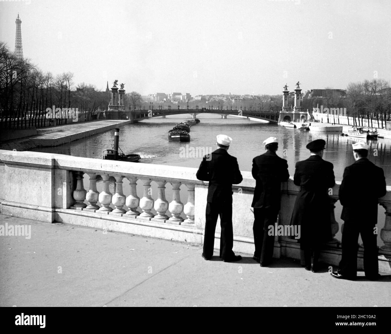 Paris Pont de la Concorde – US Navy men watch barges pass in 1945. Three navy seamen and one Chief Petty Officer stand and look west over the River Seine towards Pont Alexandre III and beyond. The tow barge is about to pass under the bridge that they are standing on while pulling at least five cargo barges behind it. The Eiffel Tower is in the left background and the Palais de Chaillot is rear center. The navy men (as well photographer Clarence Inman) are each stationed at the Paris field office of the Field Photo Branch, O.S.S. Stock Photo