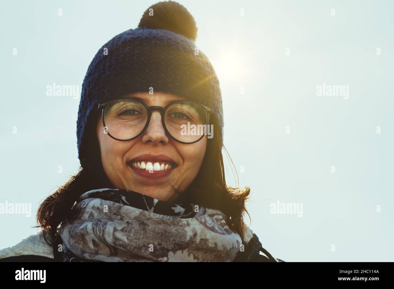 Backlit smiling woman in glasses and knitted beanie wearing a warm winter scarf in a low angle close up headshot against the sun looking down at the c Stock Photo