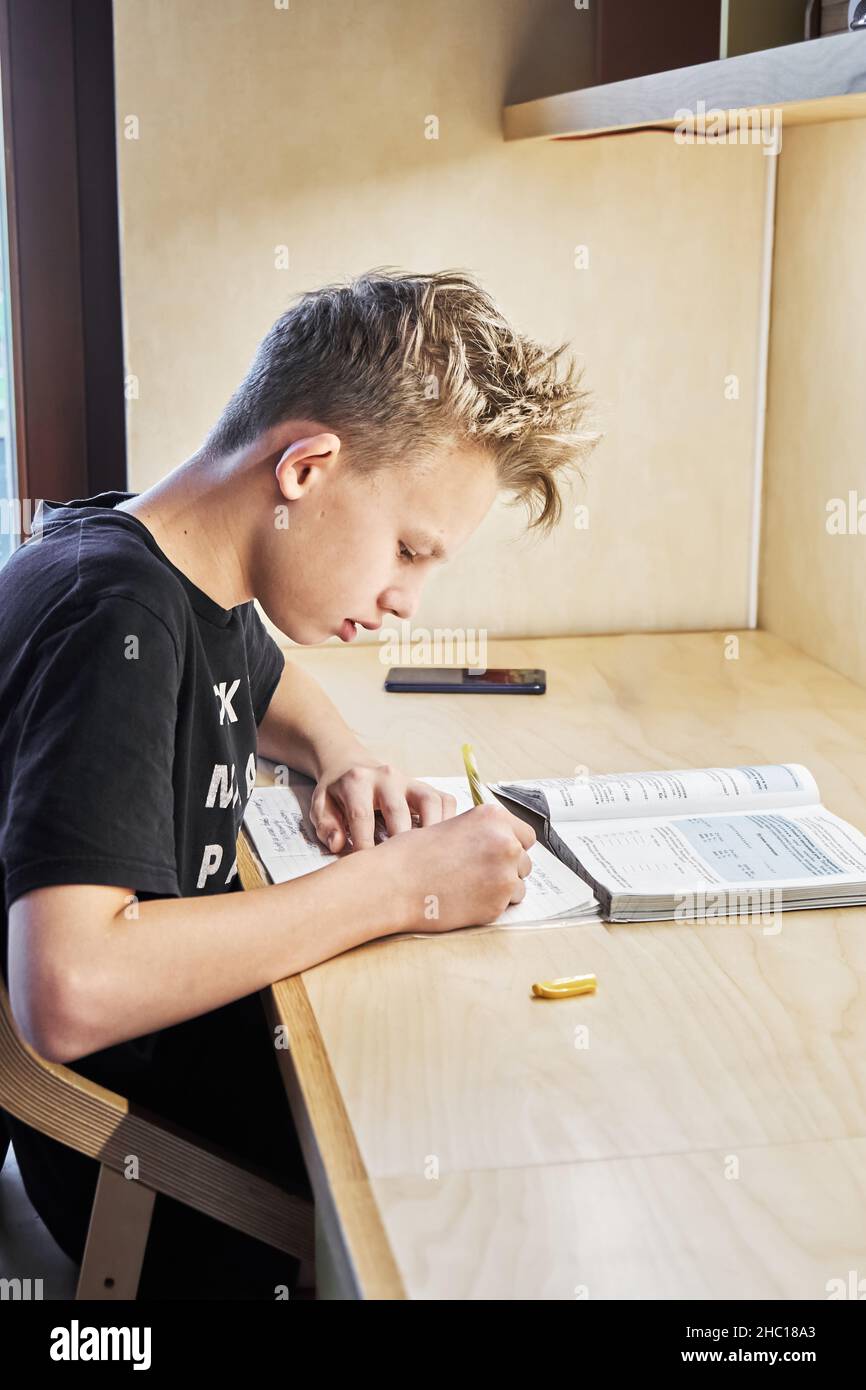 Concentrated teenage boy in black t-shirt does homework writing task in copybook at large wooden table in room close side view Stock Photo
