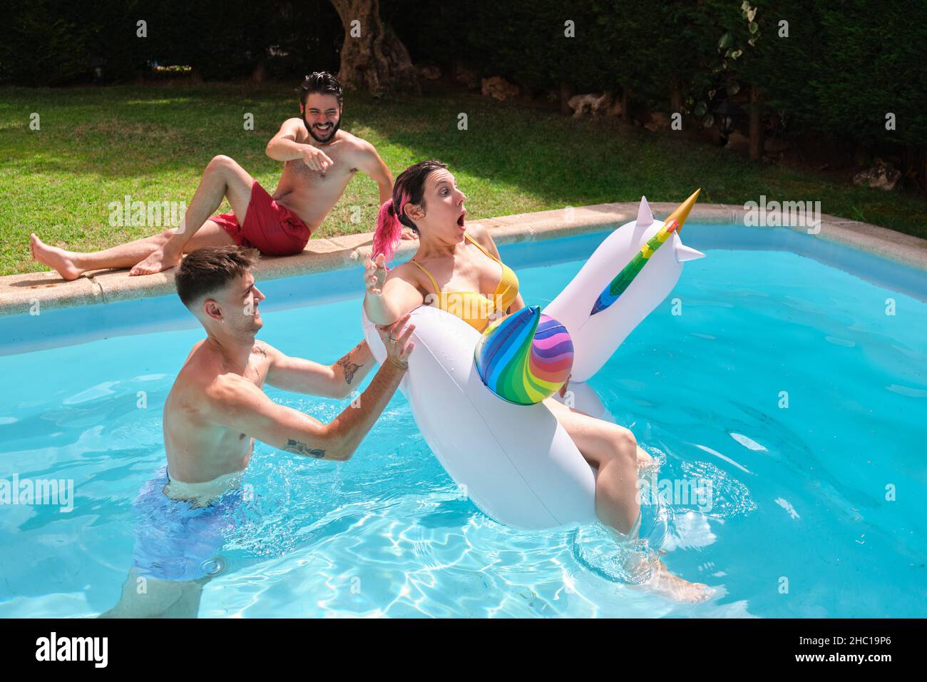 Young man throwing woman from a rainbow unicorn float into the swimming pool. Stock Photo