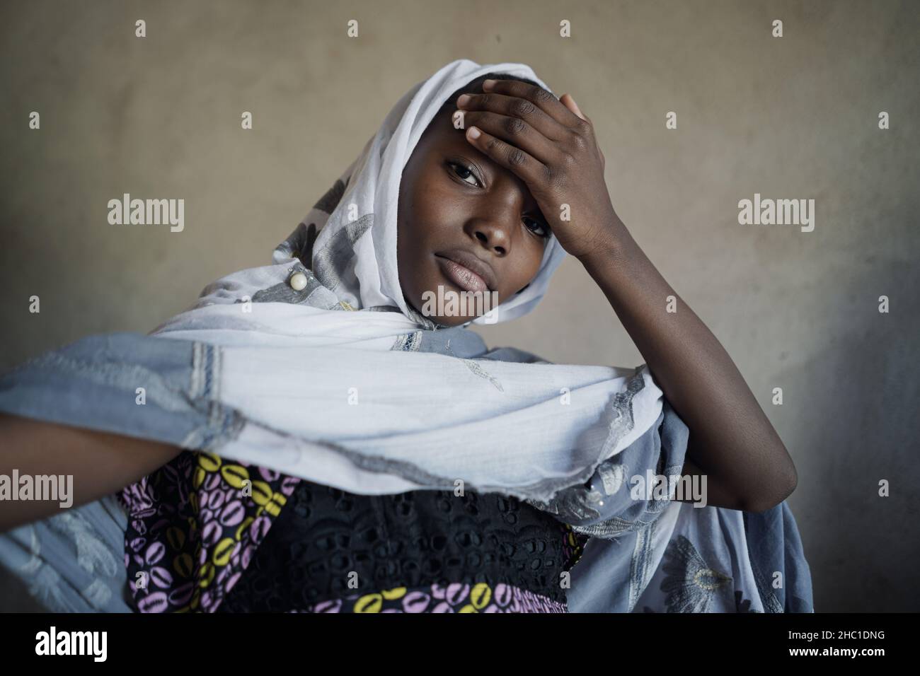 Beautiful black african teenage girl with a long and wide white scarf wrapped around her head and shoulders holding her hand on her forehead; adolesce Stock Photo