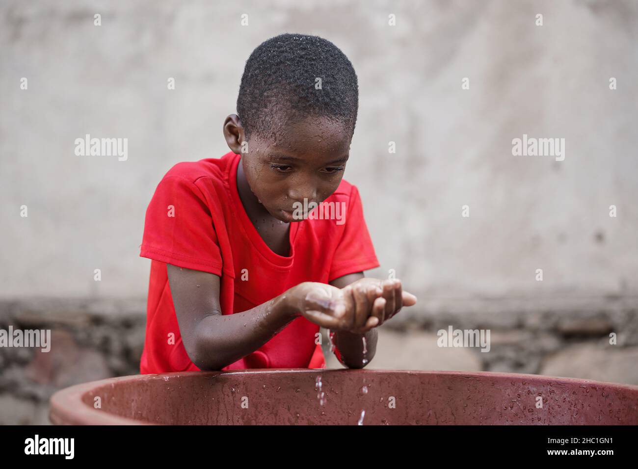Little black African boy fetching water with his cupped hands from a tub to wash his face or to drink Stock Photo
