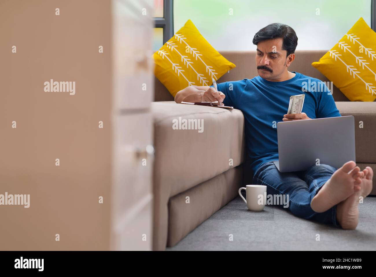 Man calculating on paper while sitting on floor with laptop on his lap Stock Photo