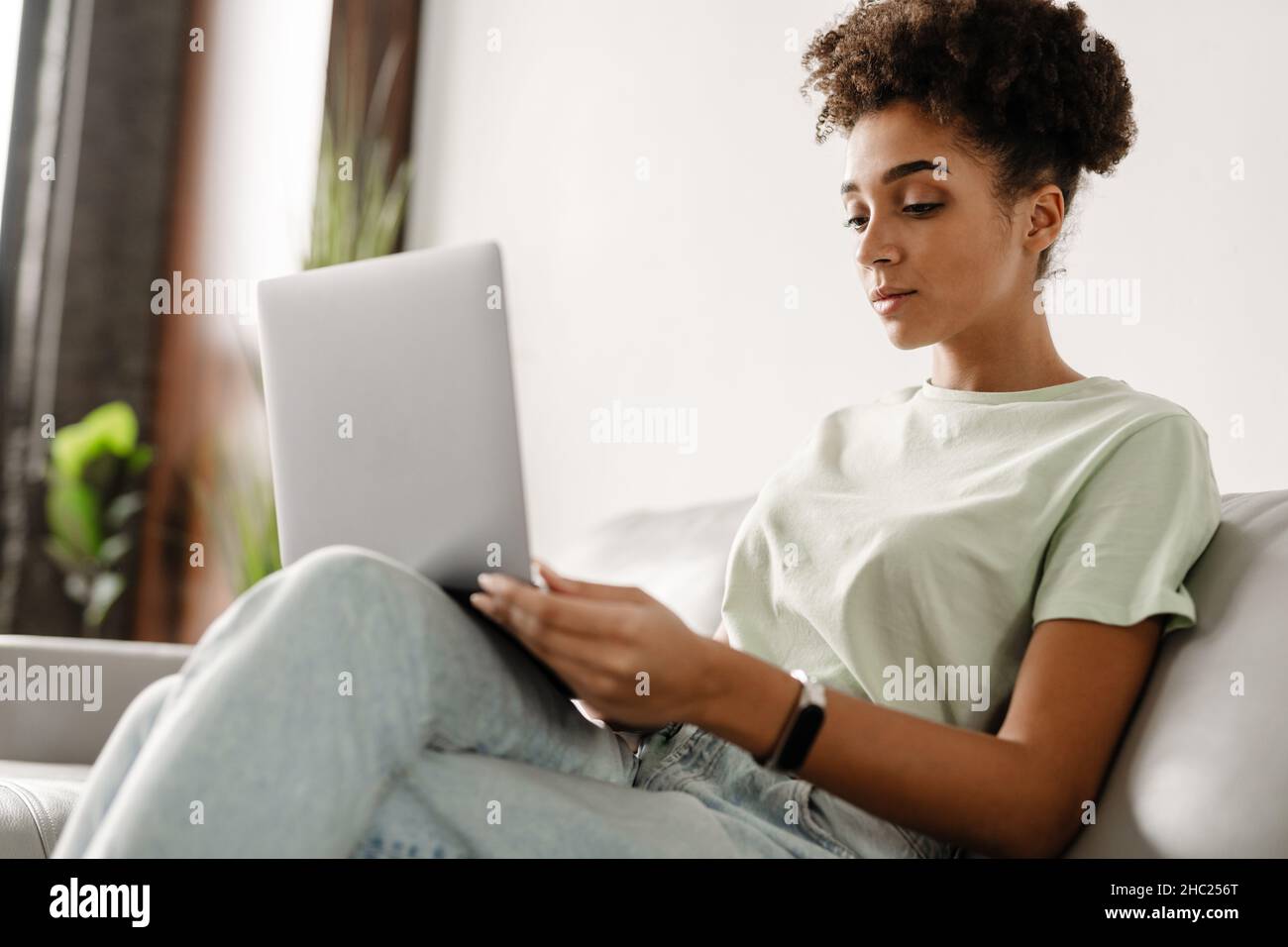 Young black woman working with laptop while sitting on couch at home ...