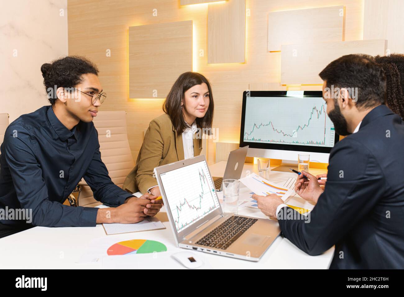 Millennial colleagues sitting at office desk, looking at computer and talking about business issues. Diverse employees communicate near each other and negotiating on projects together Stock Photo