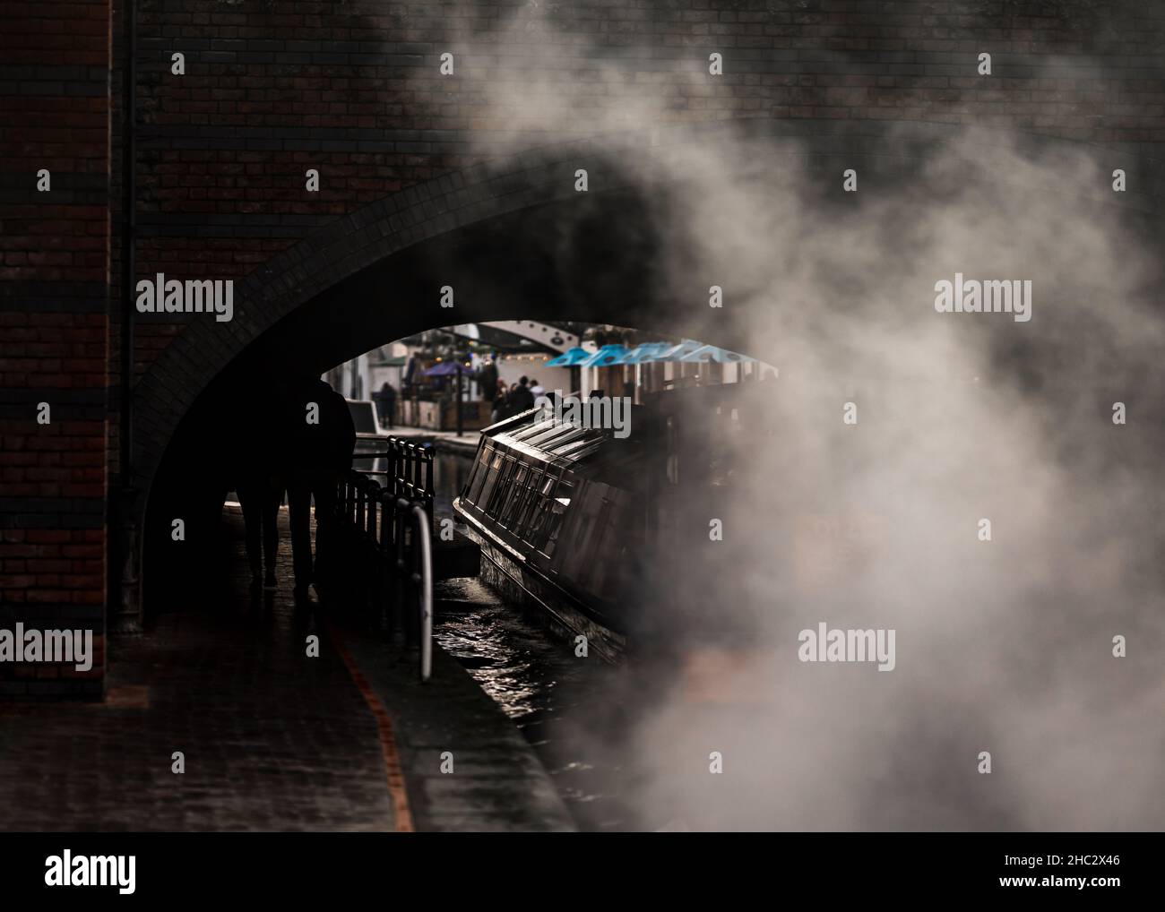 Canal riverboat passing under old arch bridge with atmospheric engine smoke from diesel powered narrow boat barge motor. Stock Photo