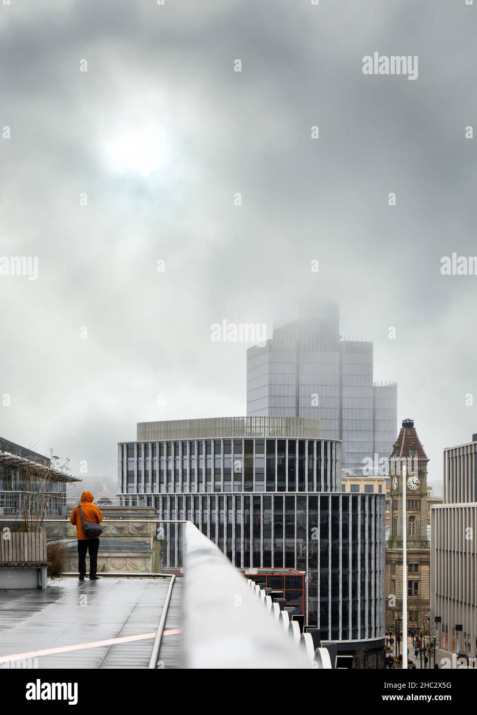 Anonymous man high up in bright coloured coat stood alone on rooftop looking out over view of city skyscrapers low cloud and stormy fog weather. Stock Photo