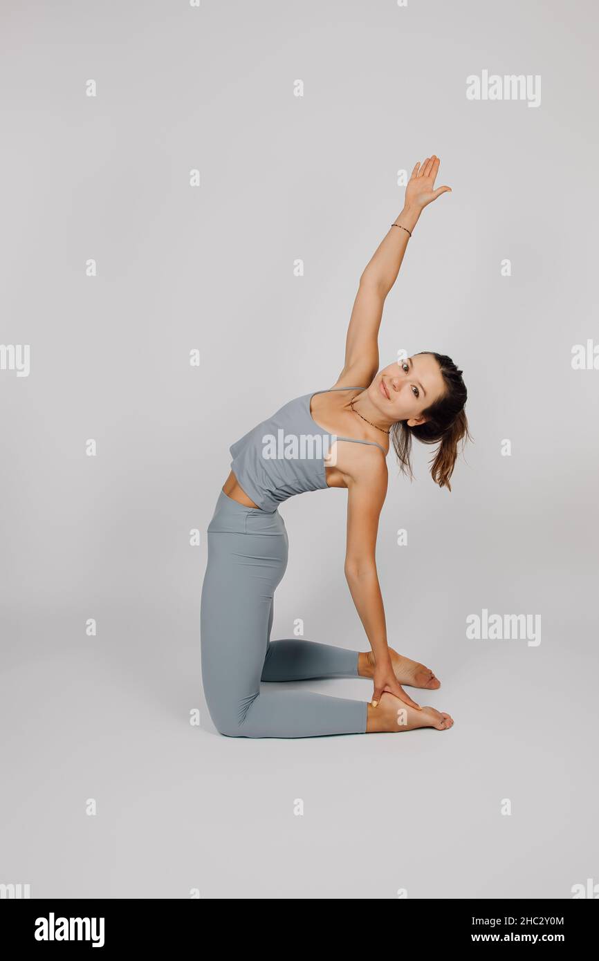 Brunette poses on isolated background. Young woman in gray tracksuit for fitness or yoga does stretching exercise, kneels and raises her hand up, side Stock Photo