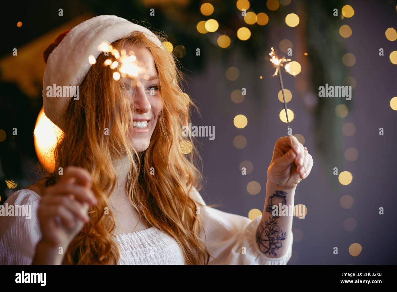 charming red-haired girl in a santa hat with sparklers in her hands smiling at the camera. Stock Photo