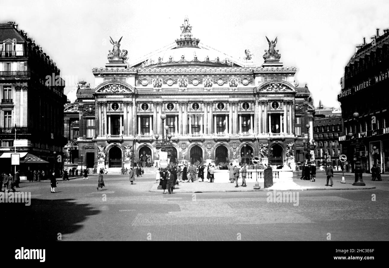 Paris Place de l'Opera wide view towards Palais Garnier, 1945. Buildings to either side are signed as the Grand Hotel and the Societe Laitiere Maggi. Dozens of people are out walking or standing near the Metro. The only vehicle in view is a partially concealed jeep. Stock Photo
