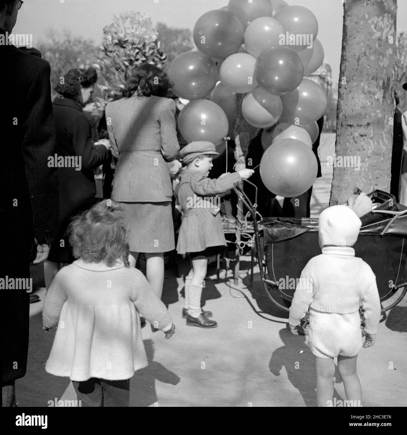 Two photographs of a scene where children are getting balloons in an unidentified Paris park in 1945. One photograph shows three parents, three children standing, and a partially obscured baby in a stroller. The other photo shows a smiling balloon-man (slightly out of focus) surrounded by several men. Some of the men wear uniforms. Stock Photo