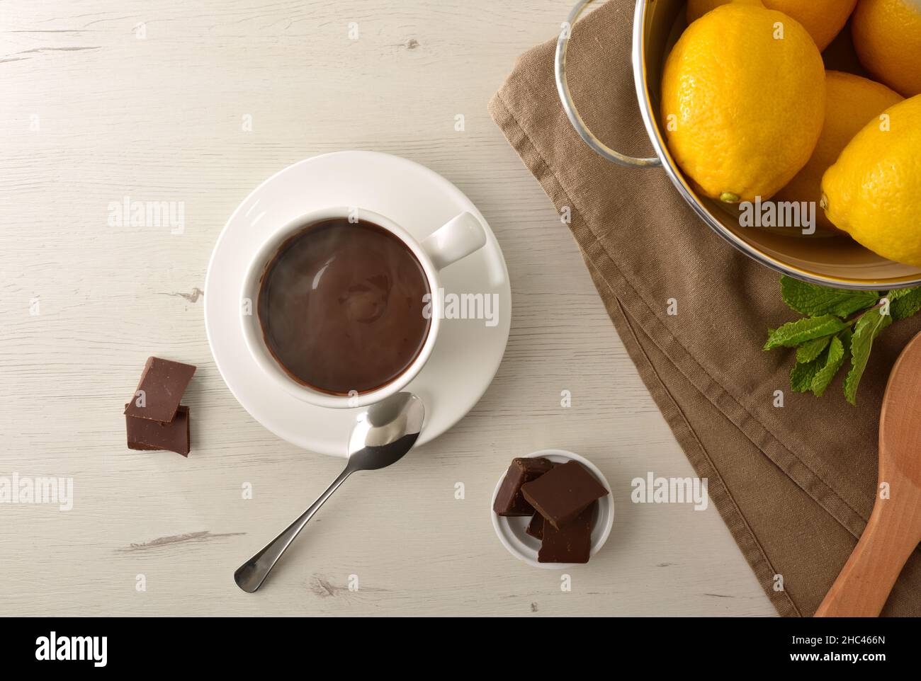 Hot chocolate in white ceramic mug, chocolate pieces in bowl on wooden table and lemons. Top view. Horizontal composition. Stock Photo