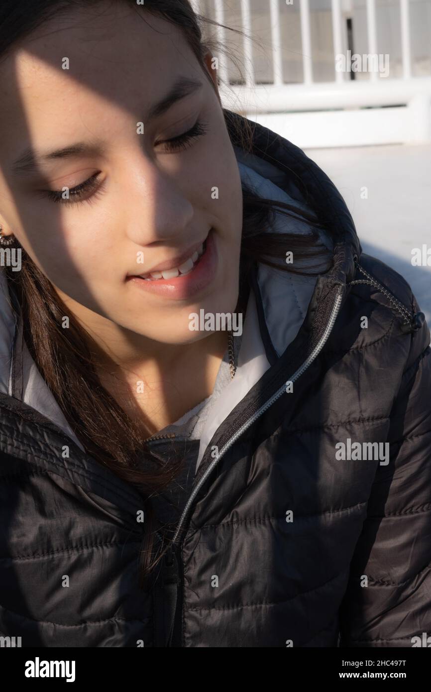 Close-up of a smiling teenage girl looking down shyly. Sunlight illuminates her face which is partially covered by a side shadow. Stock Photo