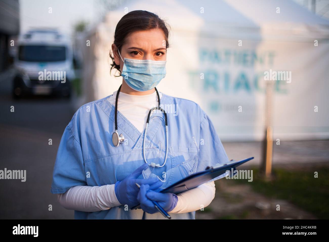 Serious female UK NHS doctor wearing protective equipment face mask,standing next to triage tent and ambulance car in front of emergency hospital,worr Stock Photo