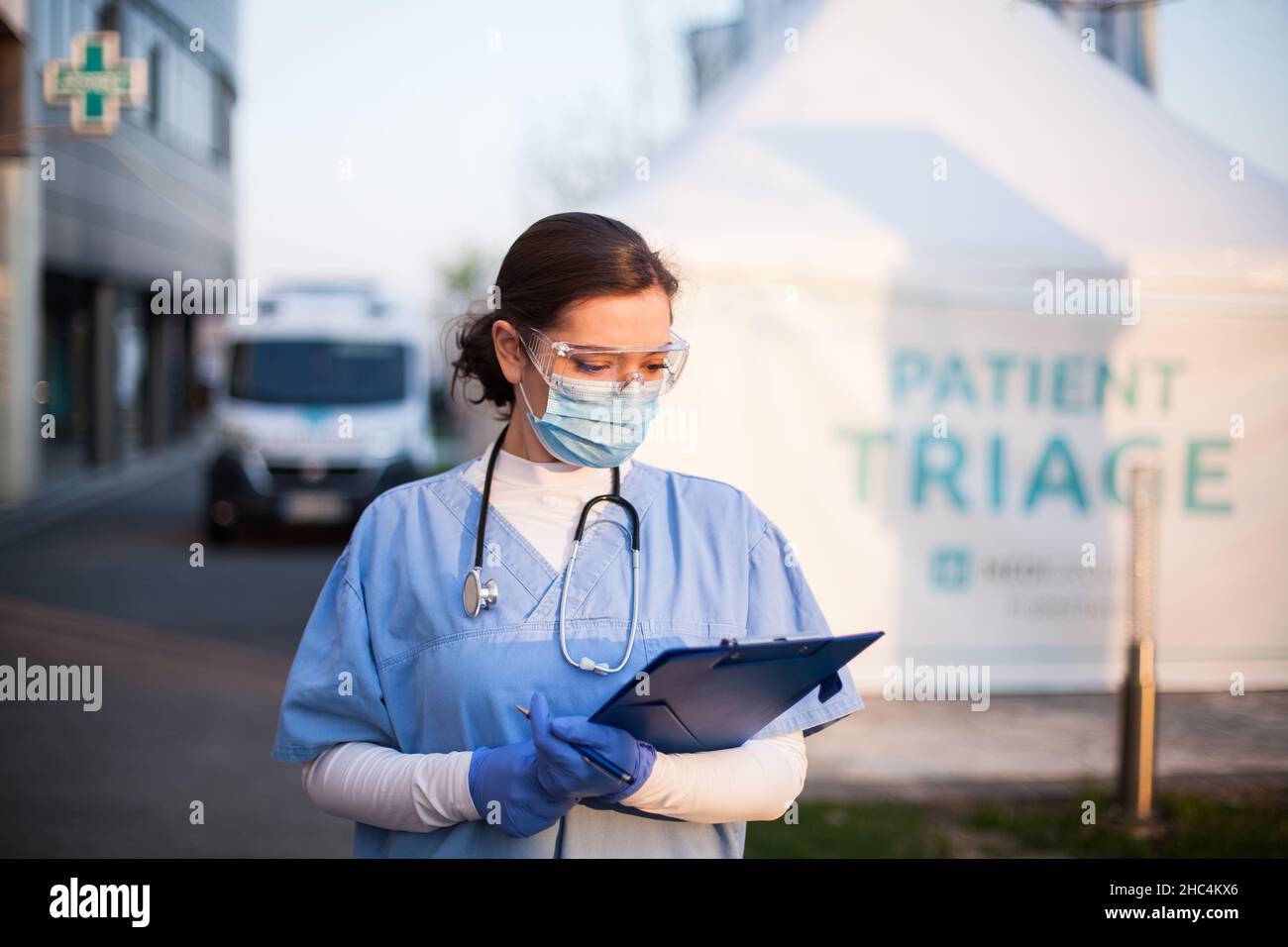 Female medical NHS doctor checking patient admission clipboard form in front of hospital,frontline first responder next to patient triage tent with am Stock Photo