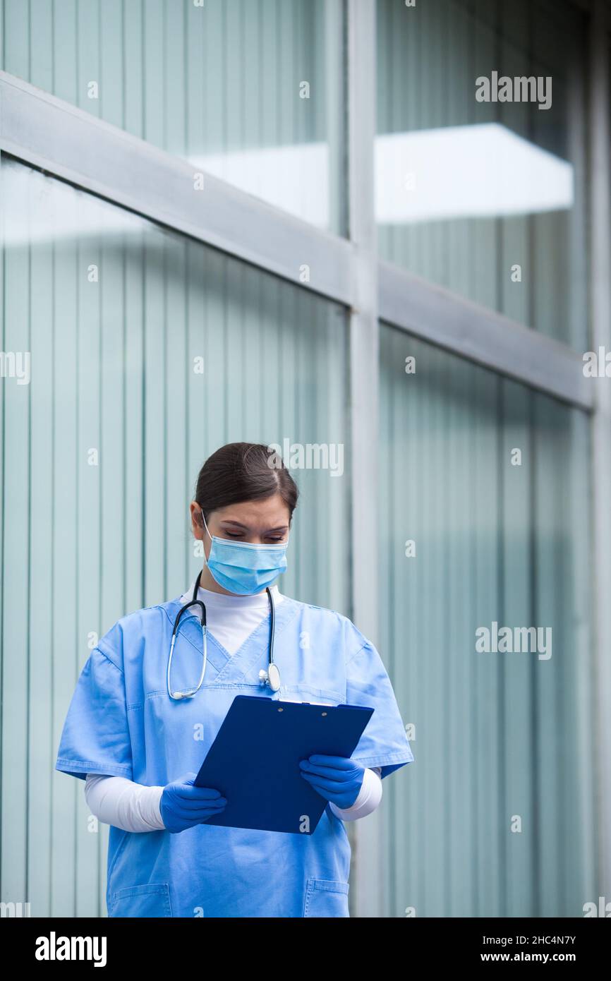Female medical doctor holding patient clipboard form,analyzing data in hospital corridor,wearing blue protective PPE scrubs:stethoscope,surgical prote Stock Photo