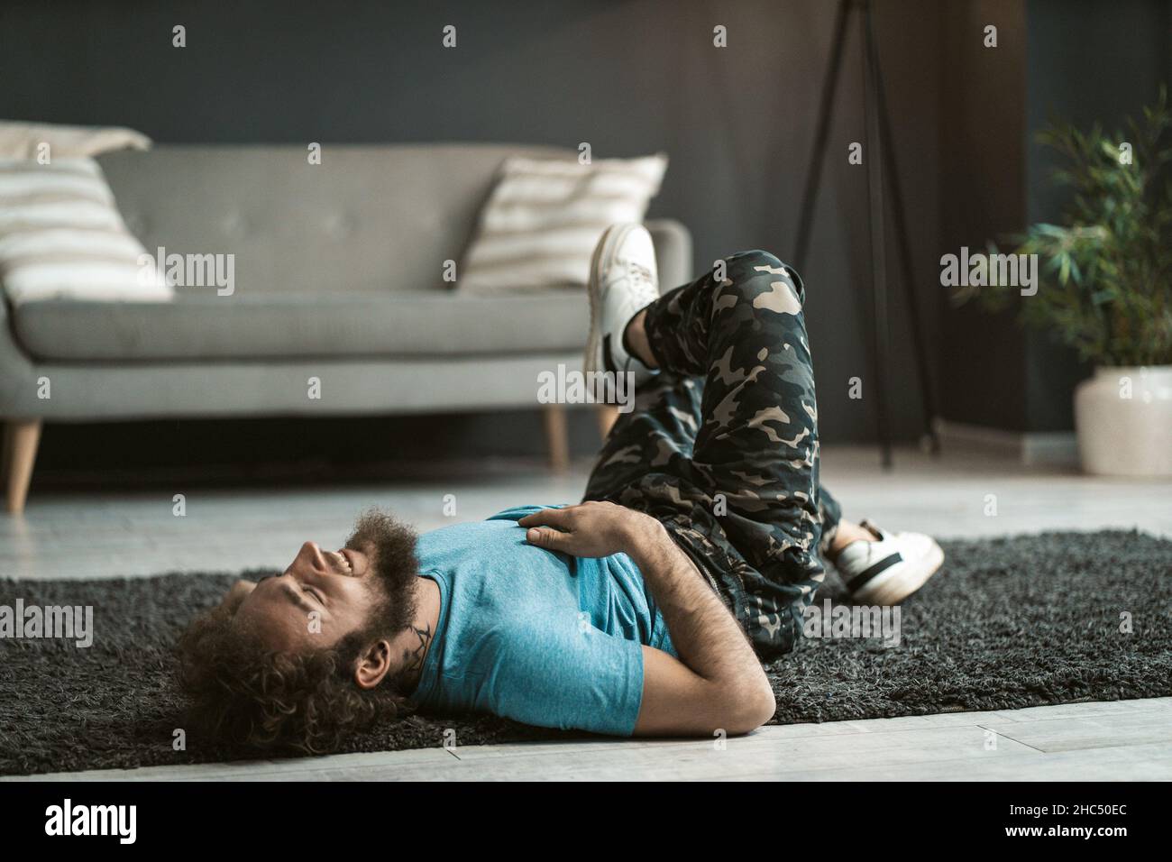 Handsome Slender Guy is having Fun on the Carpet on the Floor in the Middle of the Room. Man in a Blue T-shirt and Military Pants has a Rest on the Carpet. Gray Sofa Background. Copy space . Close-up . High quality photo Stock Photo