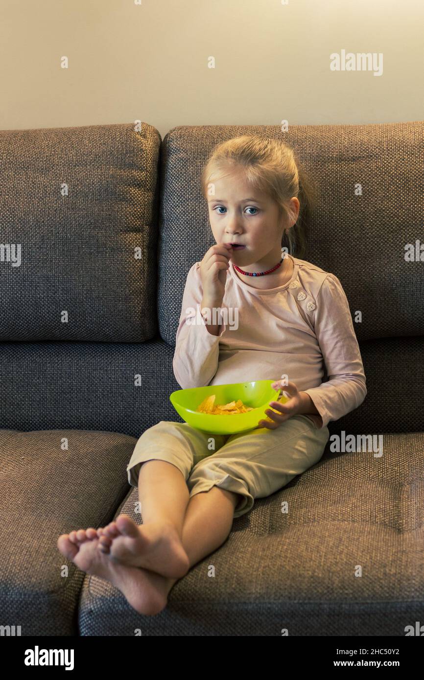 Beautiful little girl eating unhealthy food while watching TV at the sofa Stock Photo