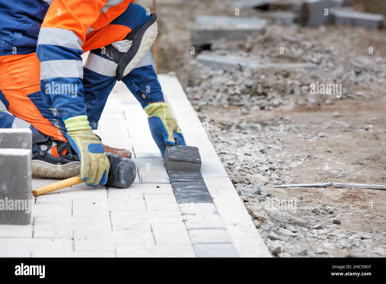 A worker in a blue and orange overalls squatting down paving paving slabs using a rubber mallet. Selective focus, copy space. Stock Photo