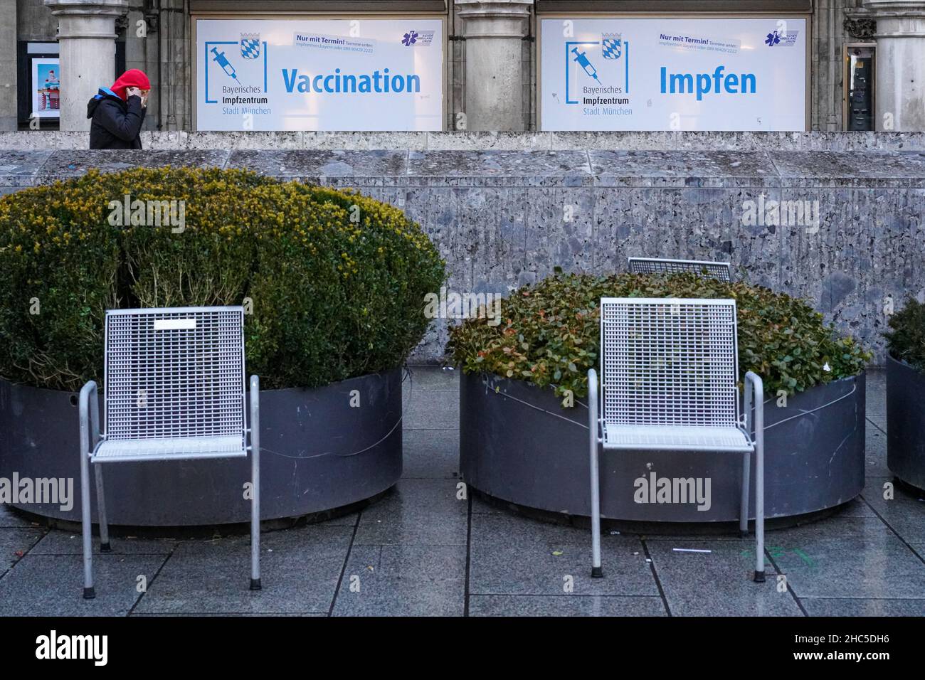 Two chairs stand on a square in front of two oversized planters. View of an advertising banner of a vaccination centre. Stock Photo