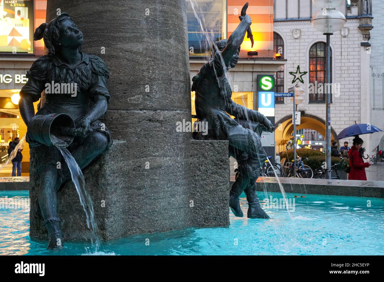 Fountain with two sculptures in the evening hours at Marienplatz in Munich with a view of the S-Bahn and U-Bahn sign of the station Marienplatz entry. Stock Photo