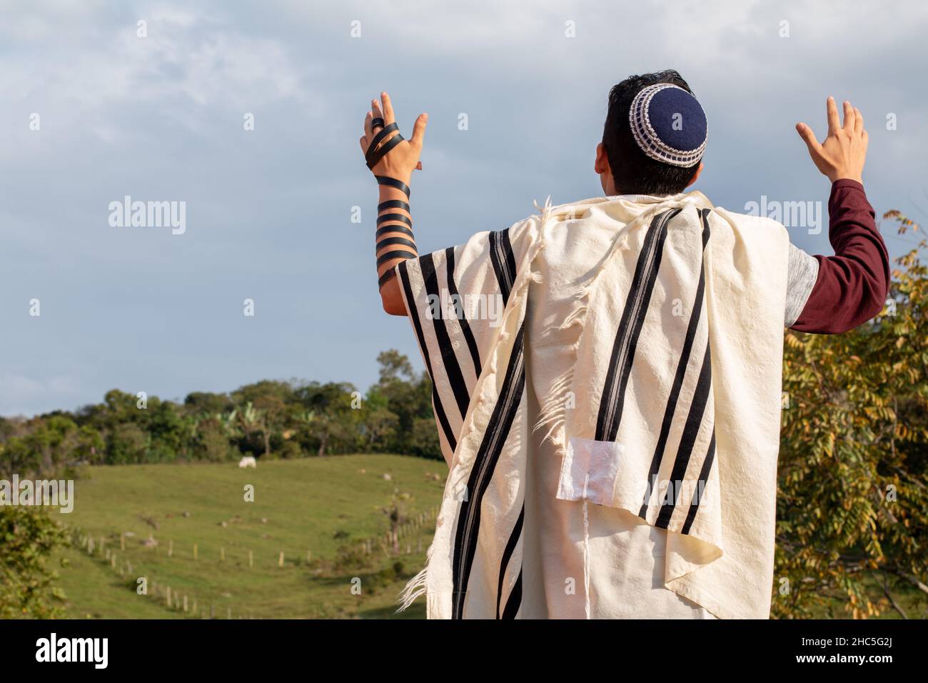 Horizontal photo of Juido Latino wearing tefilin and kippah raising his hands praying while looking at the sky with a beautiful background of natuleza Stock Photo