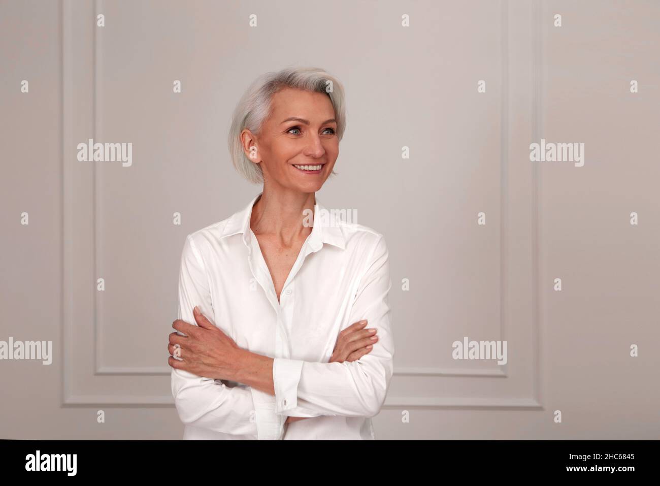 Portrait of an adult elderly self-confident woman with white hair looking awey into distance in white shirt hands crossed background wall Stock Photo