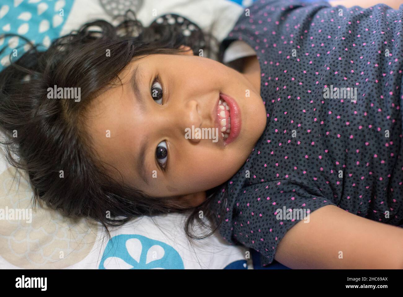 A beautiful Indonesian little girl smiling with a row of clean white teeth and long black hair on the bed, shoot from high angle Stock Photo