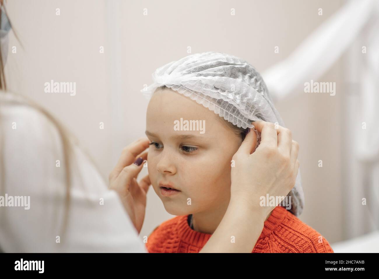 close-up of beautician hands putting white disposable medical cap on head of six-year-old girl in cosmetology office, preparing for ear piercing Stock Photo