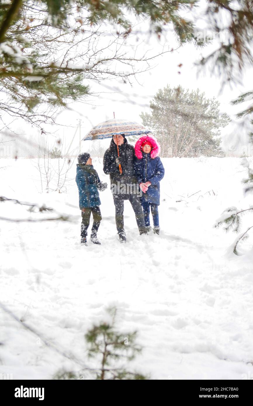 Young man in warm hat and children girl and boy on rural winter snowy background holding umbrella. Happy family, cold weather. Time together. Vacation Stock Photo