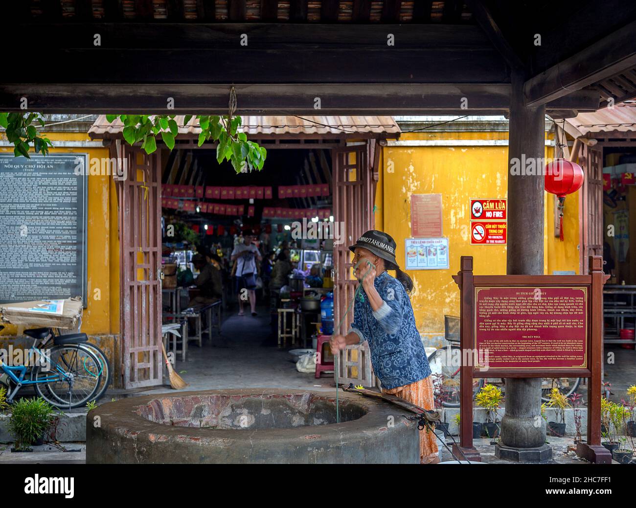 A Vietnamese lady pulls the bucket up from the well water in front of the entrance. This is in front of Central Market, Old Town, Hoi An. Stock Photo