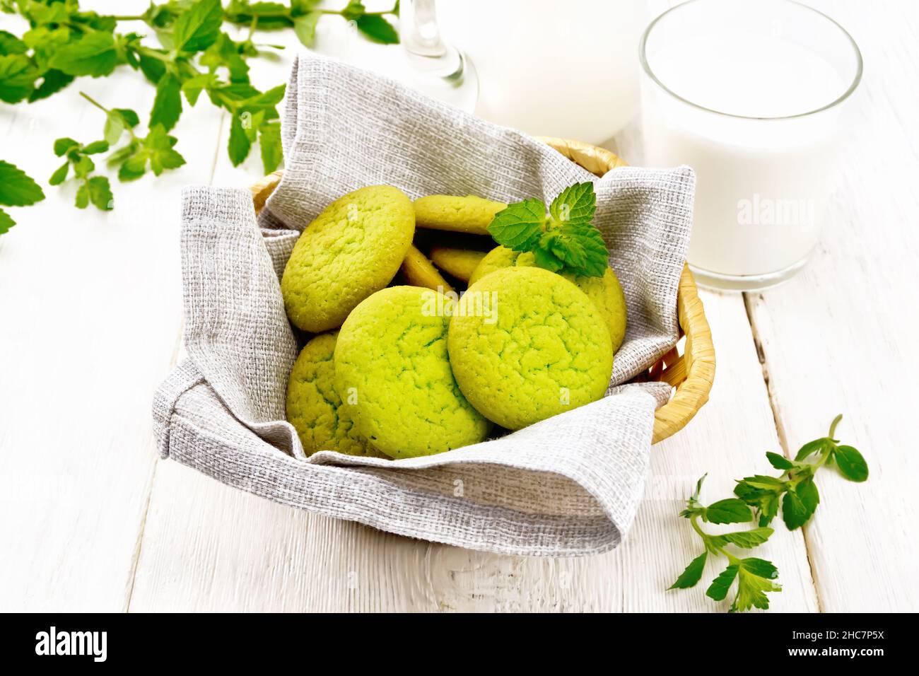 Green mint cookies on napkin in wicker basket, milk in a glass and a jug on the background of light wooden board Stock Photo