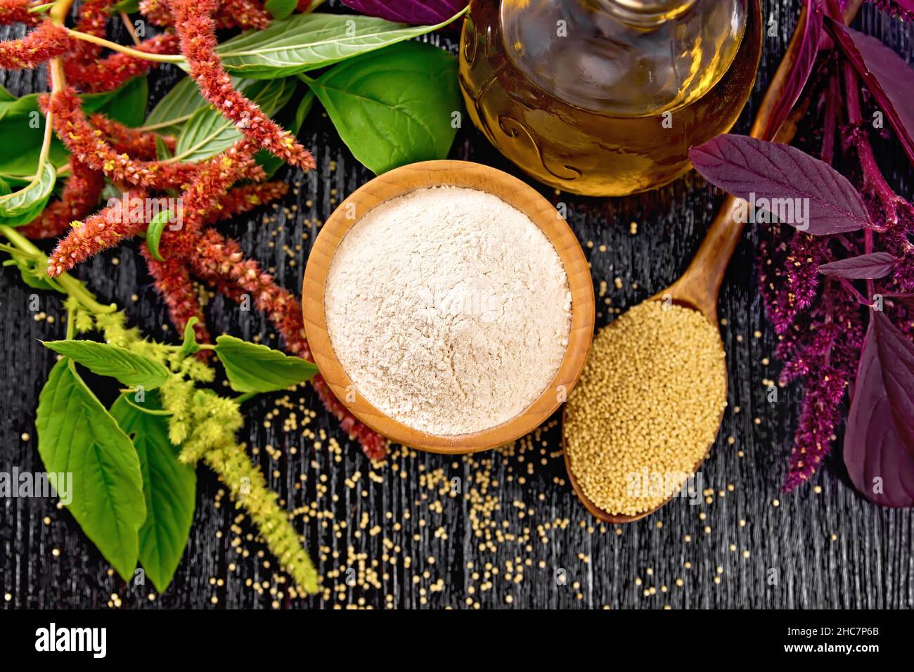 Amaranth flour in a bowl, seeds in a spoon and oil in decanter, brown, green and purple flowers of plant on black wooden board background from above Stock Photo