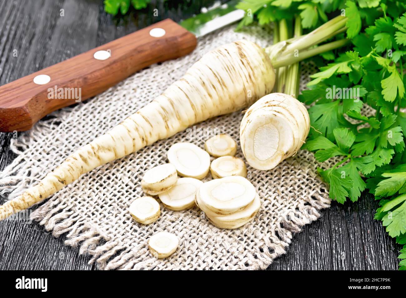 Parsley roots whole and chopped with green tops on a burlap napkin and a knife on dark wooden board background Stock Photo