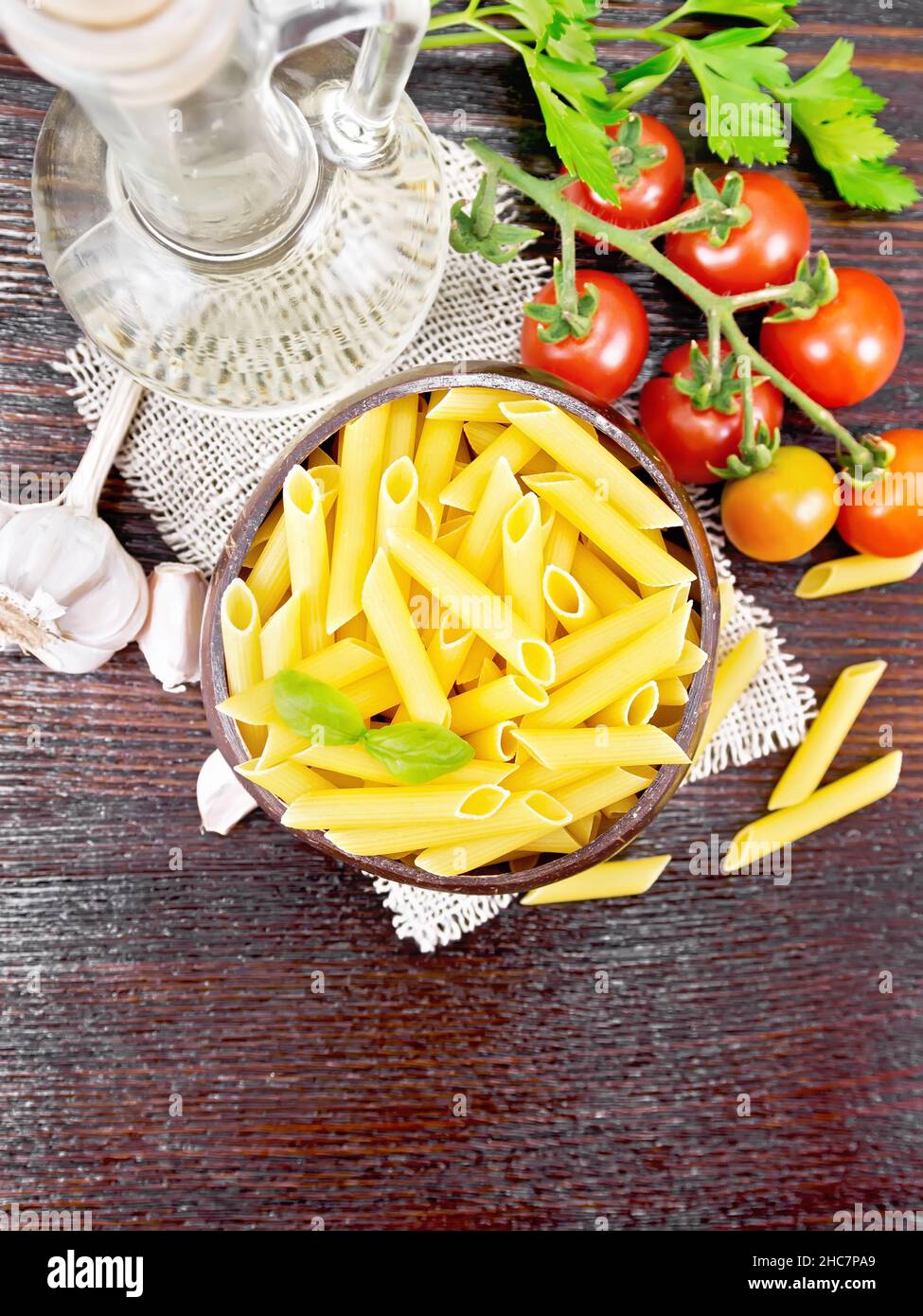 Wheat flour penne paste in a bowl of coconut shells on sacking, tomatoes, garlic, vegetable oil in a glass decanter and parsley on a wooden board back Stock Photo
